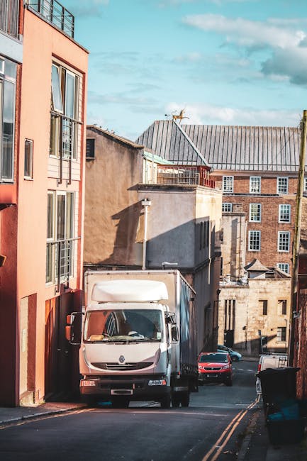 A white delivery van parked on a narrow urban street next to a multi-storey residential building with large windows and modern architecture, adjacent to older brick and concrete buildings. The van is 