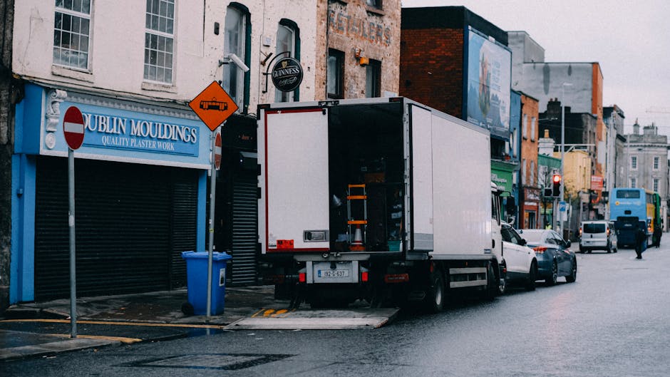 A white delivery van parked on a narrow urban street next to a multi-storey residential building with large windows and modern architecture, adjacent to older brick and concrete buildings. The van is 
